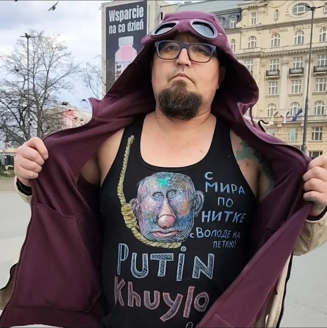 Homem barbudo de óculos e gorro com óculos de aviador na cabeça, abrindo o casaco para mostrar uma camiseta preta com a imagem de Putin enforcado e a frase Putin Khuylo em russo. Ao fundo, um outdoor polonês e um prédio antigo; O artista russo Robert Kuzovkov. -