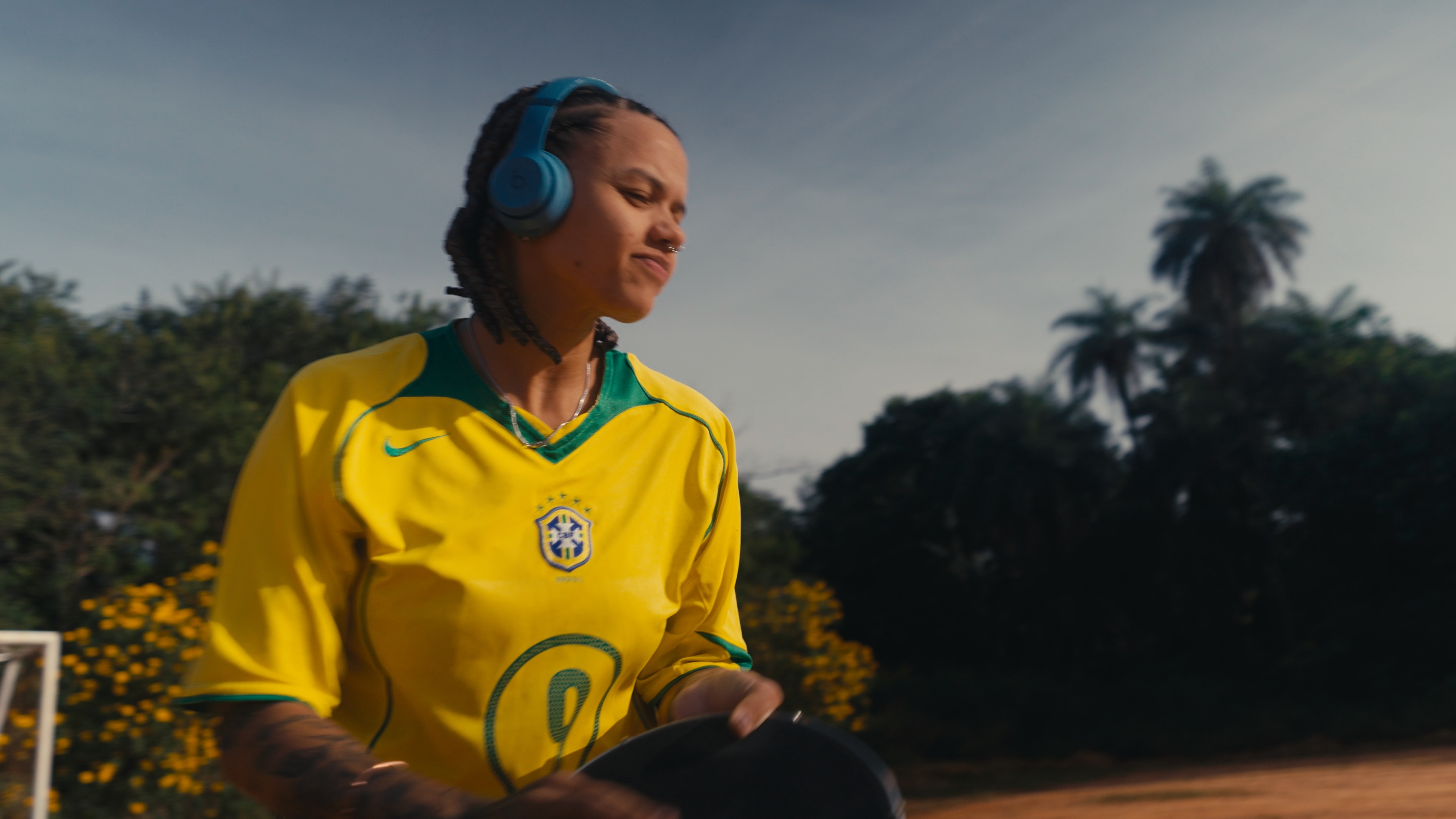 Jovem mulher de pele parda, com tranças e fones de ouvido azuis, veste camisa amarela da seleção brasileira, sorrindo levemente. Ela está em um campo de terra batida, com árvores e um céu claro ao fundo