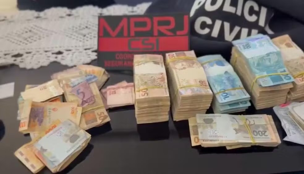 Várias pilhas de dinheiro em notas de real, incluindo cédulas de 5, 10, 50, 100 e 200, sobre uma superfície escura. Ao fundo, uma placa preta com MPRJ CSI em vermelho e parte de uma camiseta preta com POLÍCIA CIVIL em branco