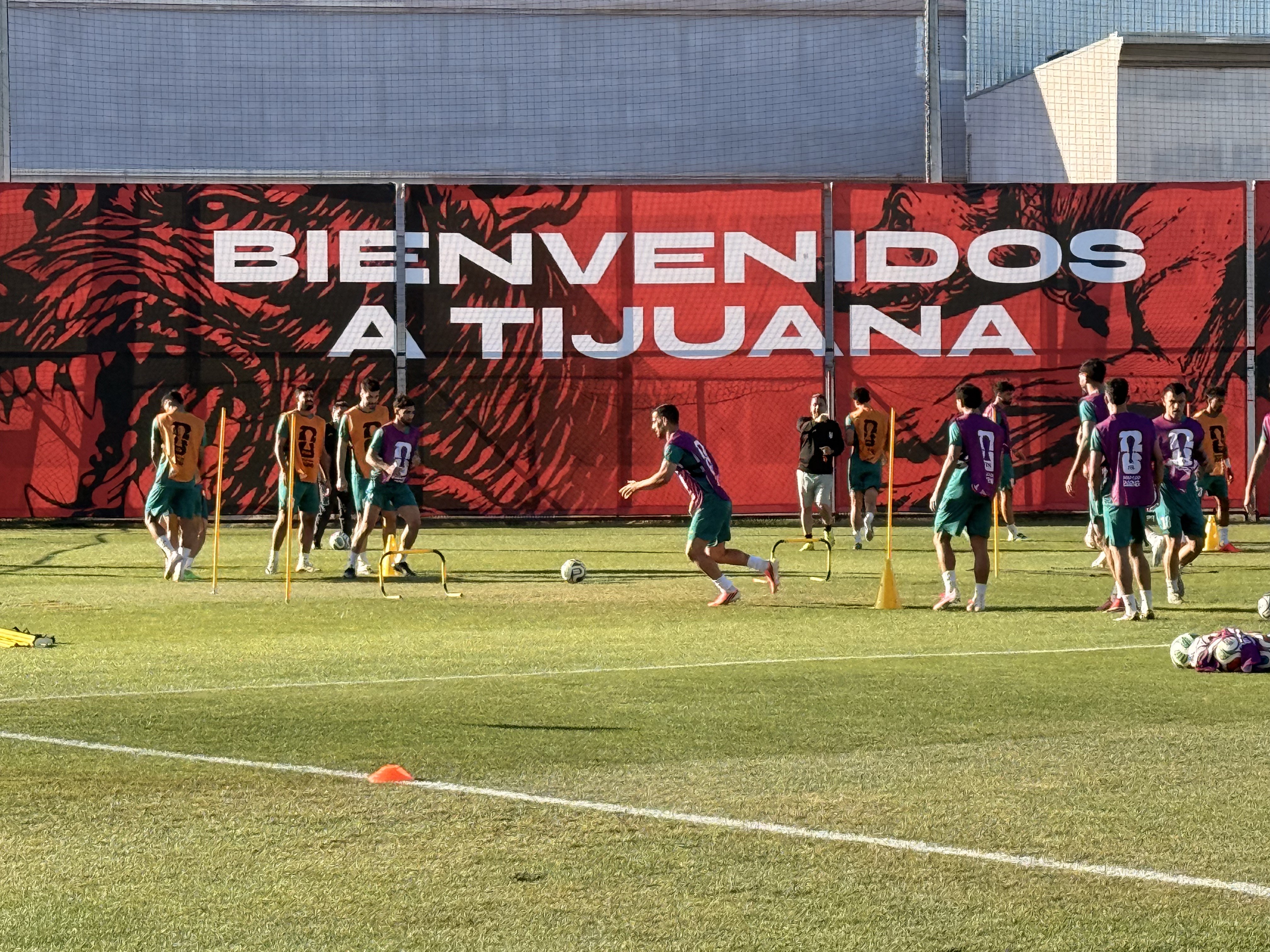 Jogadores de futebol em campo de grama verde, treinando sob um banner vermelho com a frase BIENVENIDOS A TIJUANA em branco. Um jogador de camisa roxa e calção verde corre atrás da bola, enquanto outros observam ou se preparam para o exercício. Há cones e pequenos arcos amarelos espalhados pelo campo. Ao fundo, edifícios modernos sob um céu claro