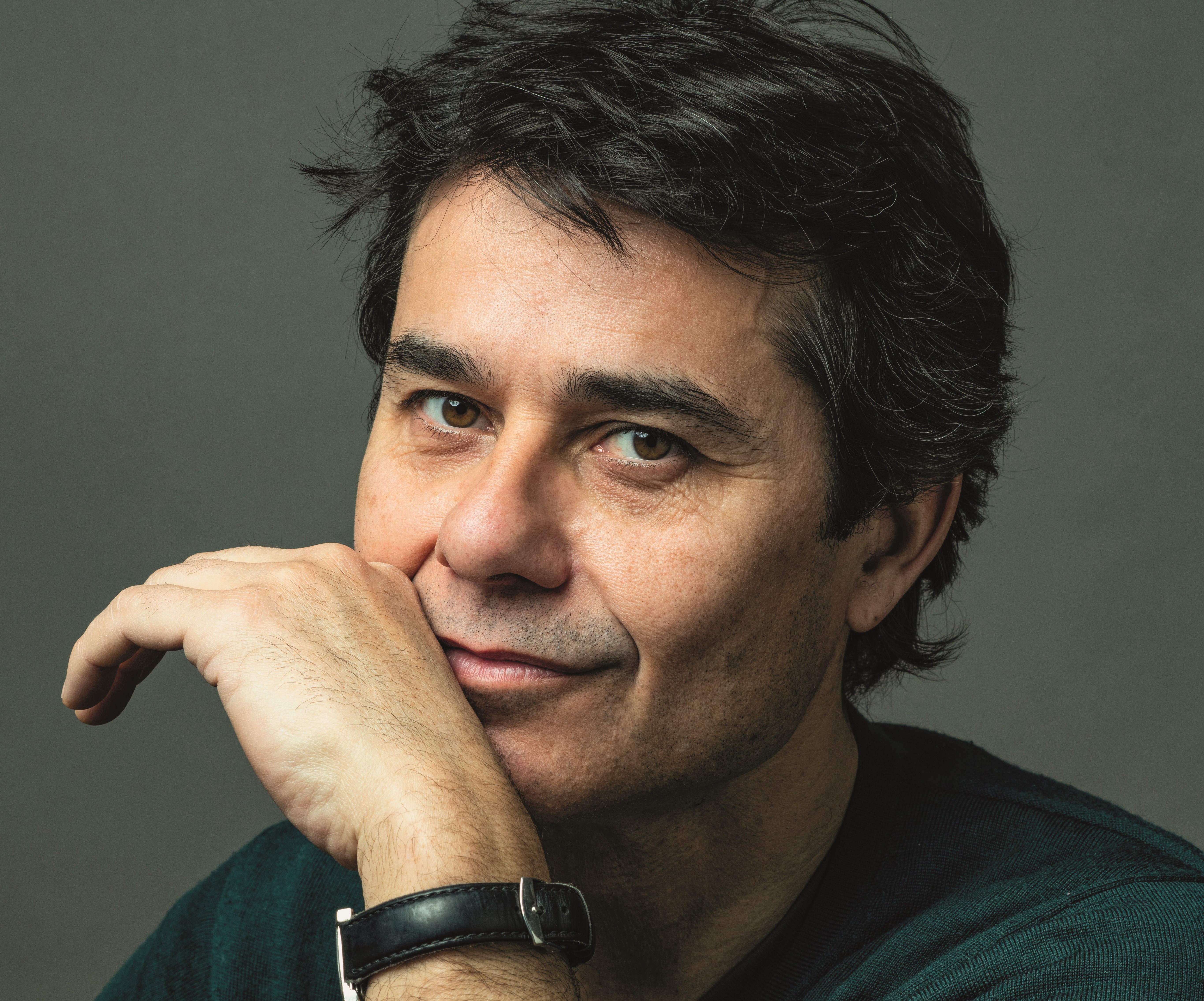 Homem de pele clara, cabelo escuro e bagunçado, sorri levemente com a mão direita apoiando o queixo, usando uma camisa escura e relógio de pulseira preta