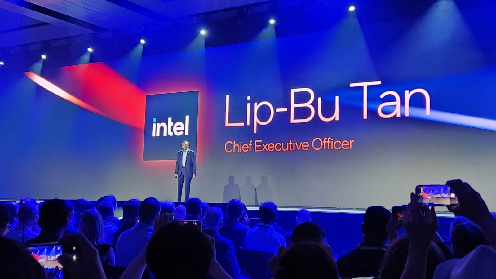 Homem de terno escuro em palco azul iluminado, com logo Intel e nome "Lip-Bu Tan, Chief Executive Officer" projetados. Plateia escura assiste, alguns filmam com celulares