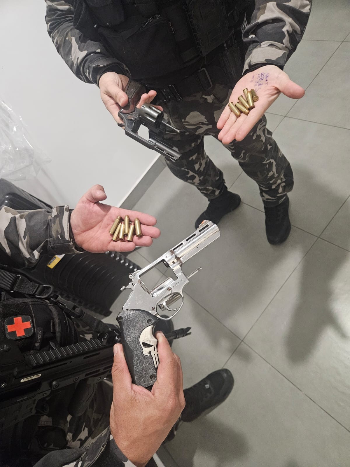Duas pessoas, vestindo uniformes camuflados e coletes táticos, exibem armas e munições. Uma segura um revólver prateado com cabo preto decorado com uma caveira branca, e na outra mão, cartuchos de munição. A segunda pessoa segura um revólver escuro e também mostra cartuchos na palma da mão. O cenário é um piso claro, possivelmente um ambiente interno.