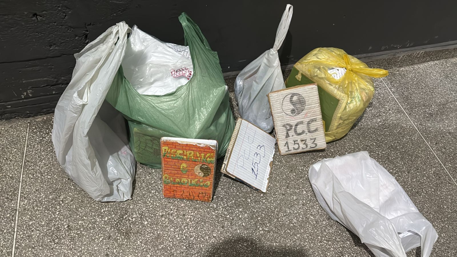 Várias sacolas plásticas de cores variadas no chão, com dois tijolos de drogas e um caderno aberto com o número 1533 escrito, e uma placa de madeira com o símbolo do yin-yang e a inscrição PCC 1533