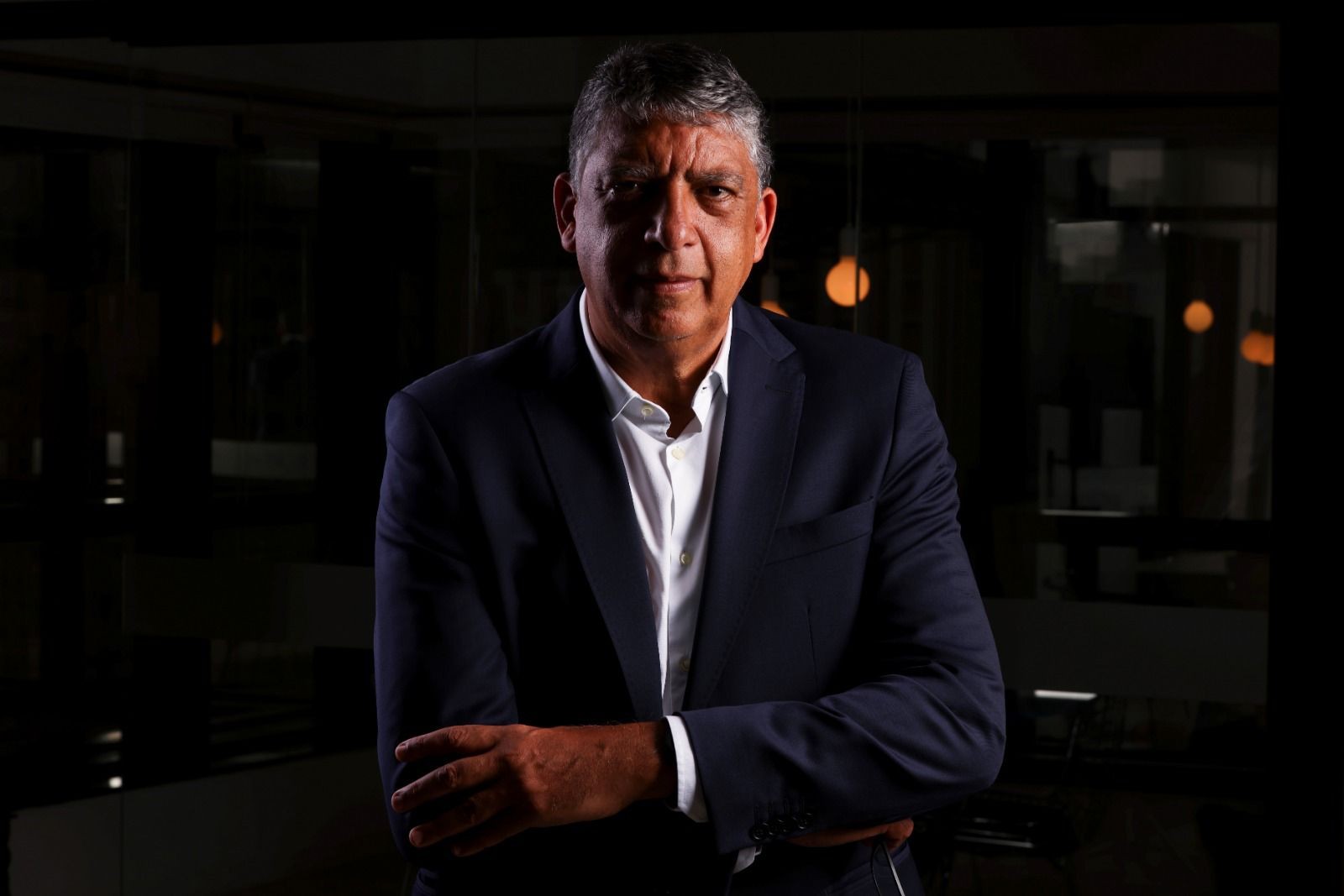 Homem de pele morena, cabelos grisalhos curtos, vestindo terno azul-marinho e camisa branca, braços cruzados, olhando sério para frente, em ambiente escuro com luzes desfocadas ao fundo