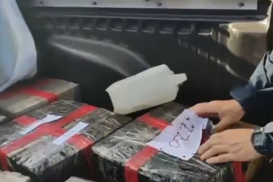 Mãos de um homem com relógio seguram um papel com "0220" sobre pacotes retangulares escuros, amarrados com fita vermelha, no compartimento de carga de um veículo, ao lado de um recipiente branco