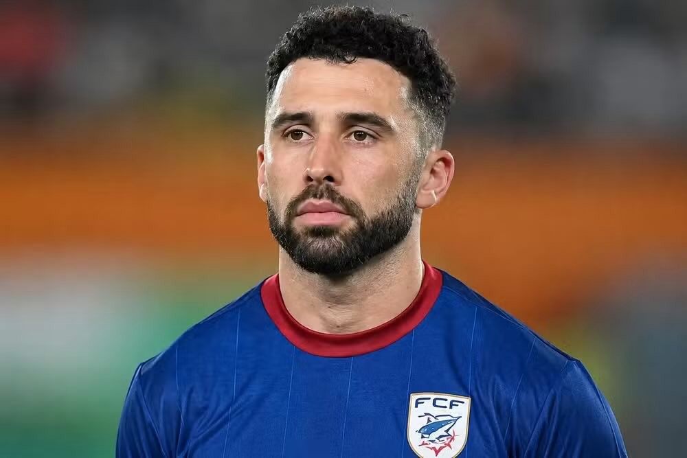 Homem de pele clara, barba e cabelo escuros, vestindo camisa azul com gola vermelha e o escudo da FCF no peito, olhando para frente com expressão séria