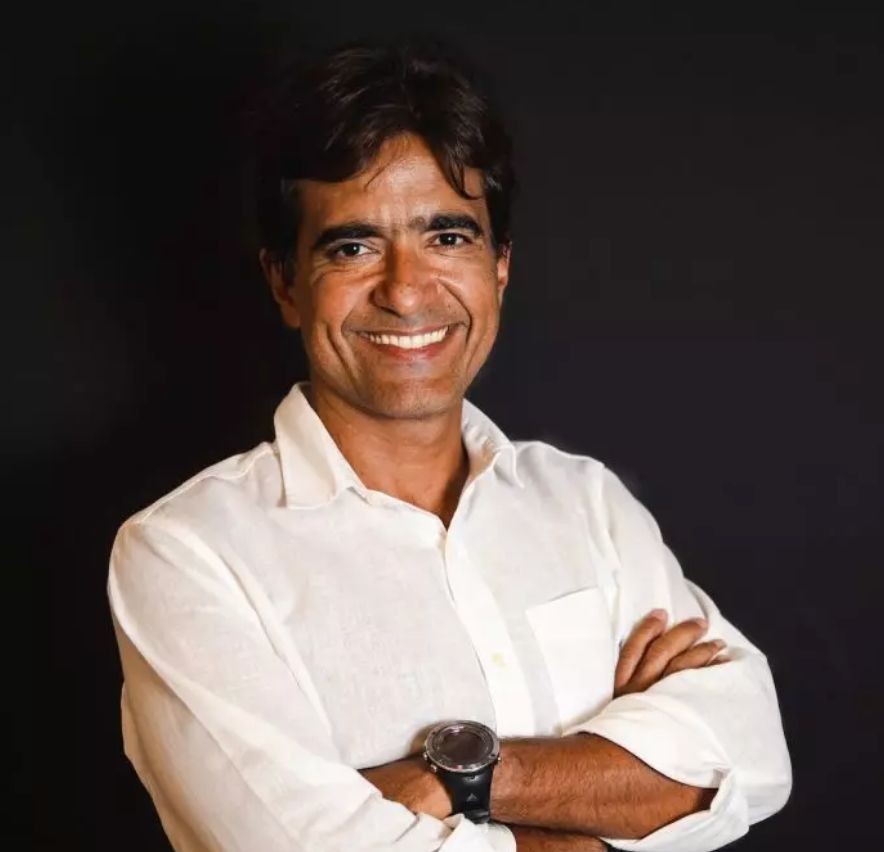 Homem de pele morena, cabelo castanho escuro e curto, sorrindo para a câmera. Veste camisa social branca de manga comprida e usa um relógio esportivo preto no pulso esquerdo. Seus braços estão cruzados na altura do peito, e o fundo é preto liso