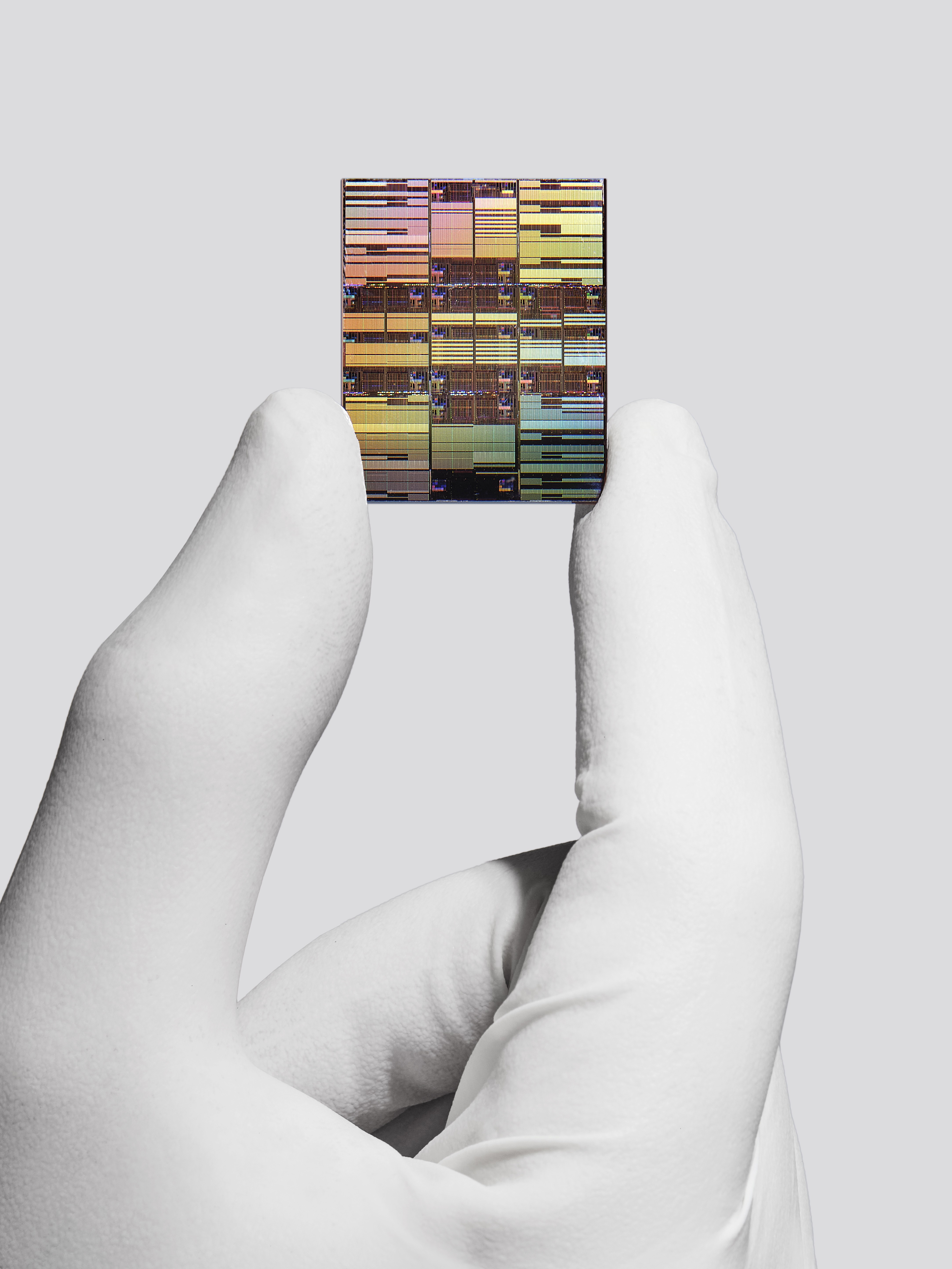Mão enluvada de branco segura um chip de computador retangular, exibindo circuitos coloridos em tons de amarelo, rosa e azul