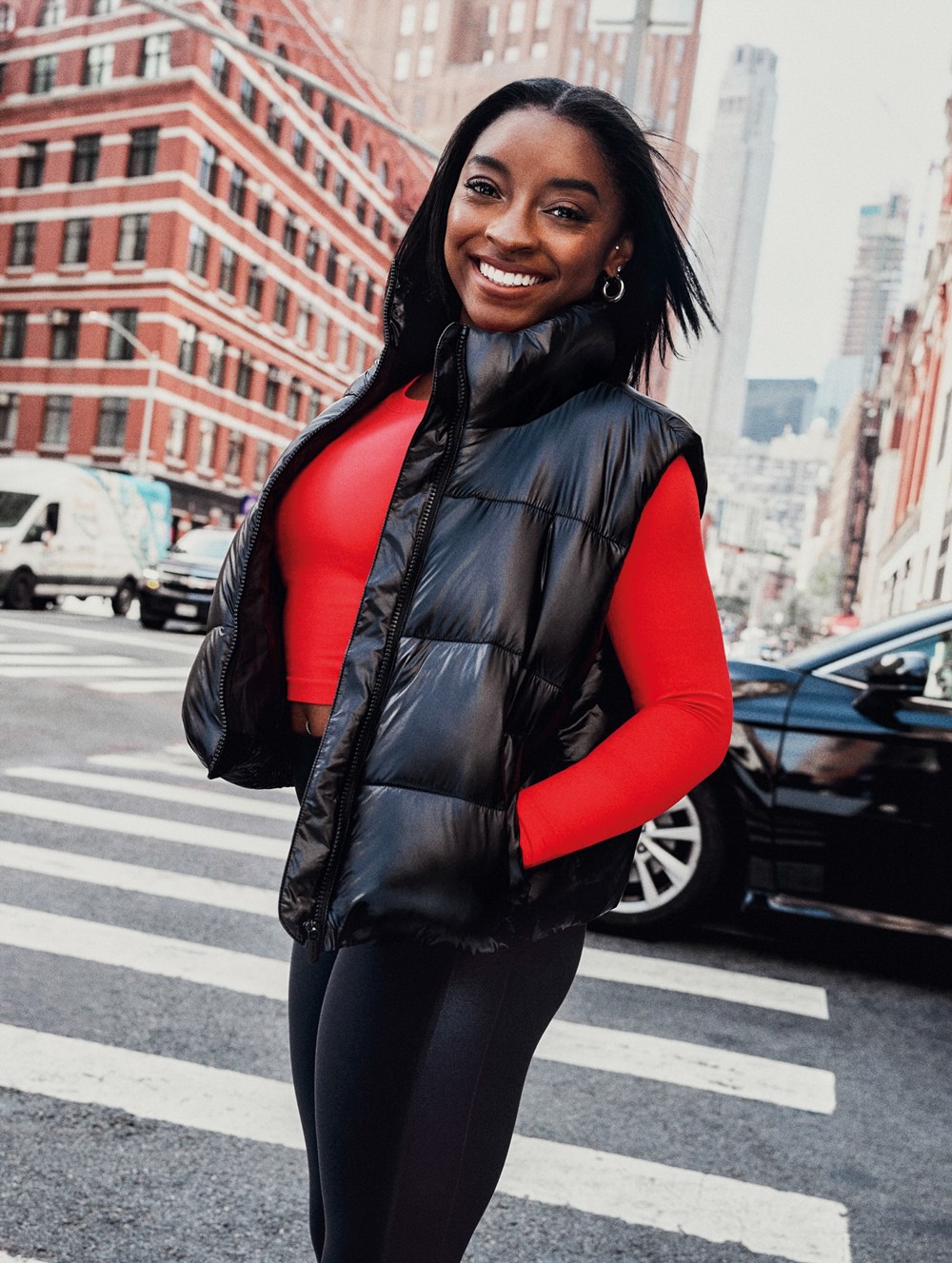 Simone Biles sorrindo, vestindo colete puffer preto, blusa vermelha de manga comprida e cal&ccedil;a legging preta, em uma rua movimentada da cidade com pr&eacute;dios e carros ao fundo