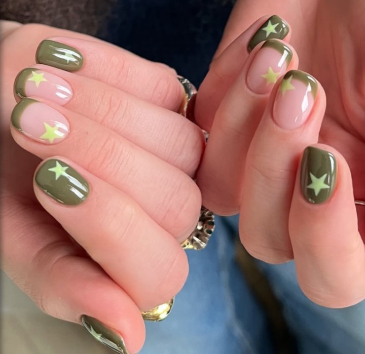 Mãos com unhas curtas e arredondadas, decoradas com esmalte verde-oliva e rosa claro, com estrelas amarelas. Algumas unhas têm a ponta verde-oliva e base rosa, outras são totalmente verde-oliva. Um anel dourado com detalhes prateados é visível no dedo anelar da mão esquerda