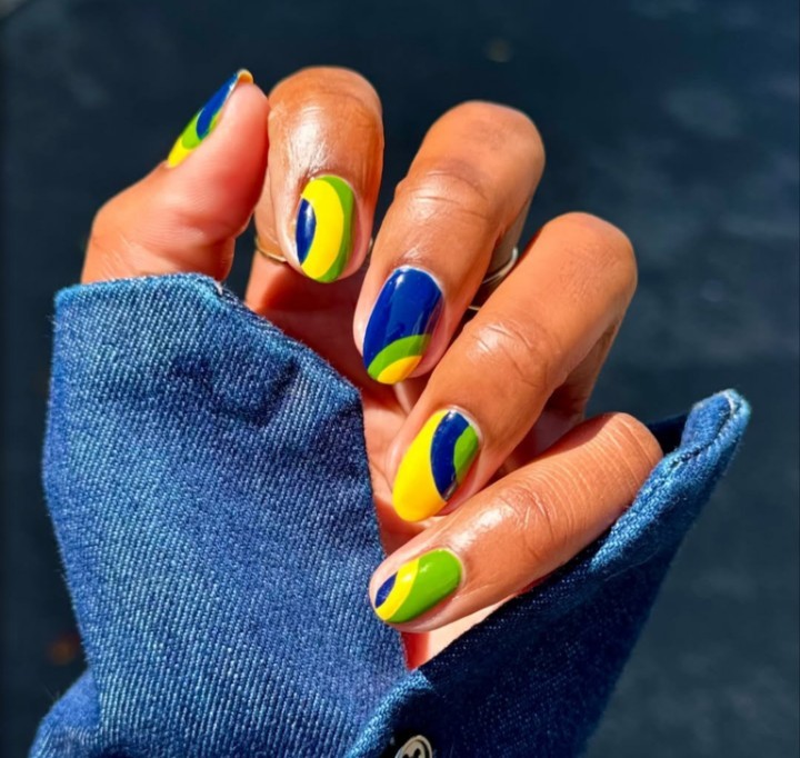 Mão de pele morena com unhas pintadas nas cores da bandeira brasileira: azul, amarelo e verde, em um fundo escuro. A pessoa veste uma manga de camisa jeans