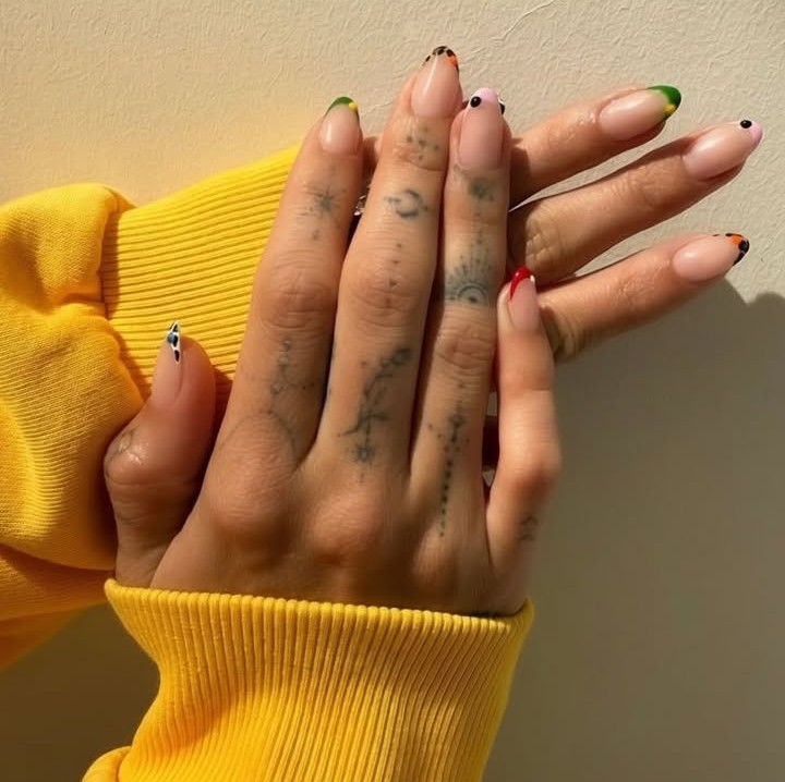 Mãos de uma pessoa com unhas amendoadas e coloridas, usando uma blusa de moletom amarela. As unhas têm pontas em tons de verde, rosa, laranja e preto, com pequenos pontos pretos em algumas. Os dedos possuem tatuagens delicadas de símbolos e constelações