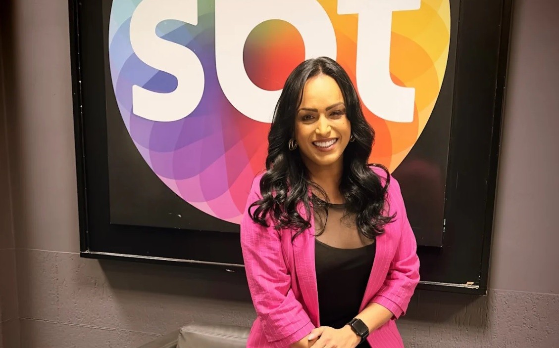 Mulher de pele morena, cabelos longos e ondulados, sorri para a câmera. Veste blazer rosa choque e blusa preta transparente no colo. Ao fundo, o logo do SBT com letras brancas sobre um círculo colorido