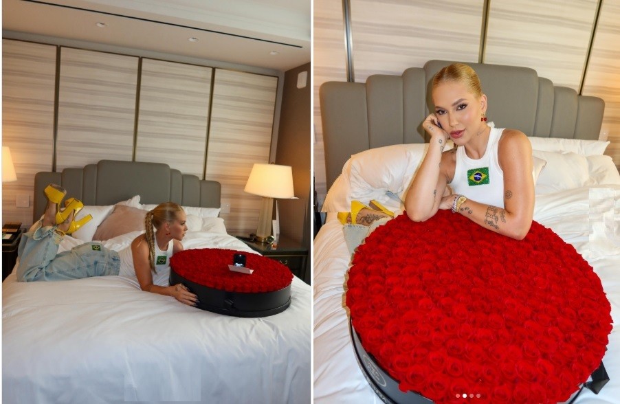 Montagem de duas fotos: à esquerda, uma mulher loira com rabo de cavalo, vestindo regata branca com bandeira do Brasil e calça jeans clara, deitada na cama com os pés para cima, ao lado de uma caixa oval preta cheia de rosas vermelhas e uma caixinha de anel. À direita, a mesma mulher, com maquiagem leve e brincos, sorri para a câmera, apoiando o rosto na mão, com a caixa de rosas em primeiro plano