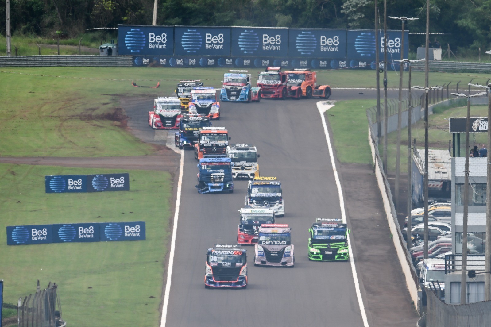 Caminhões de corrida coloridos em alta velocidade em uma pista de asfalto, com grama verde e painéis de publicidade ao redor, durante uma competição automobilística. Copa Truck