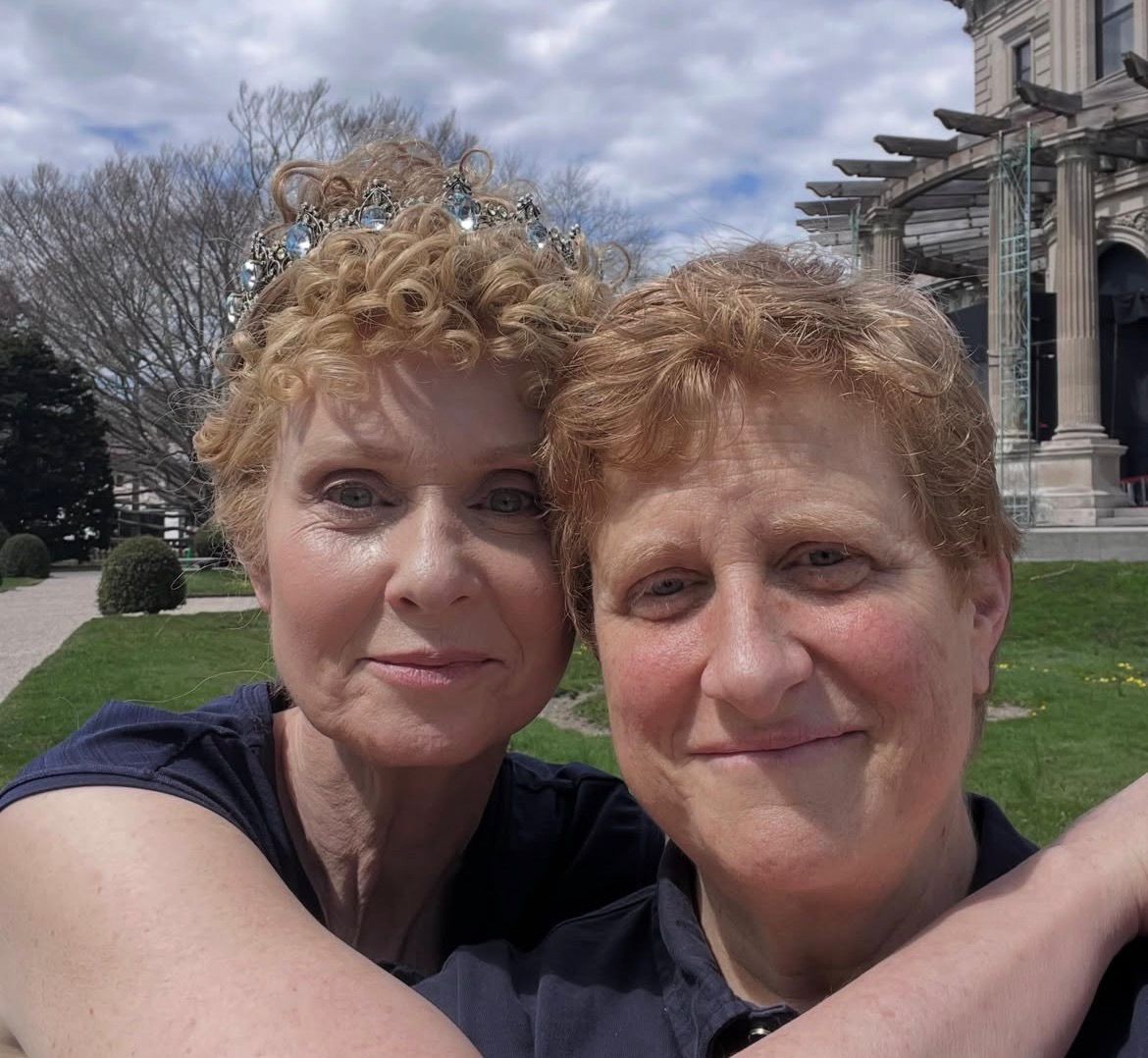 Duas mulheres sorridentes, uma com tiara de pedras azuis no cabelo cacheado loiro, abraçadas em selfie com um casarão antigo e jardim ao fundo sob céu nublado