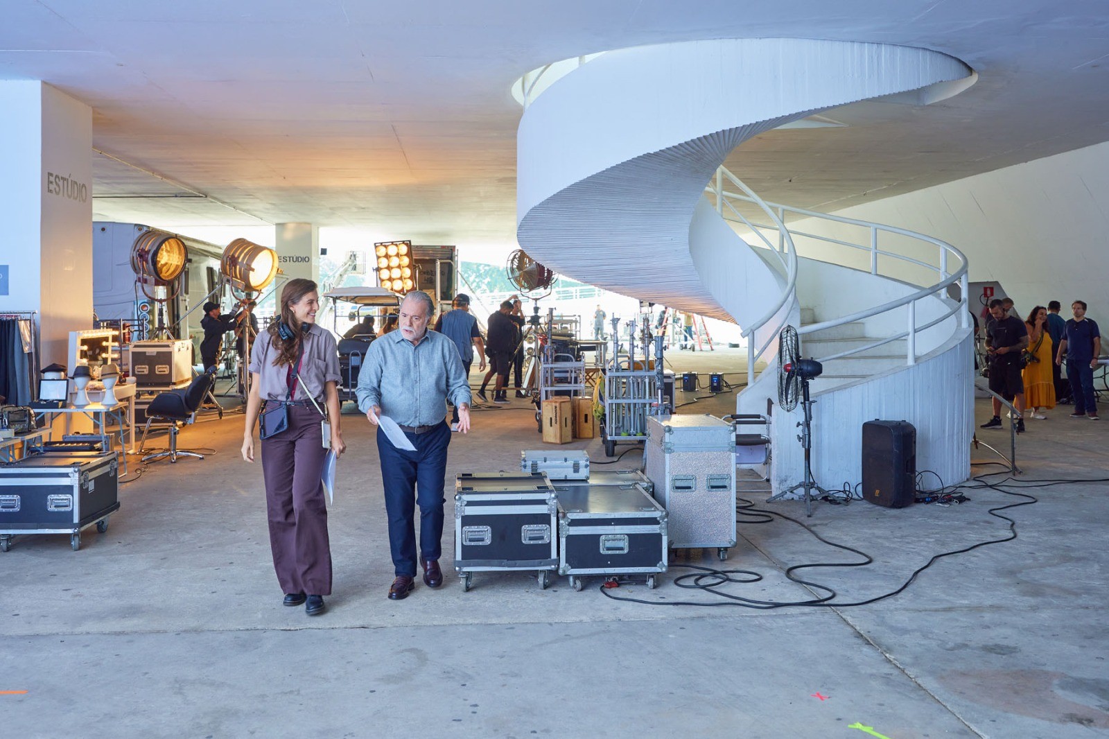 Uma mulher e um homem caminham em um estúdio de filmagem com equipamentos de luz e caixas de transporte. Ao fundo, uma escada espiral branca e pessoas da equipe.