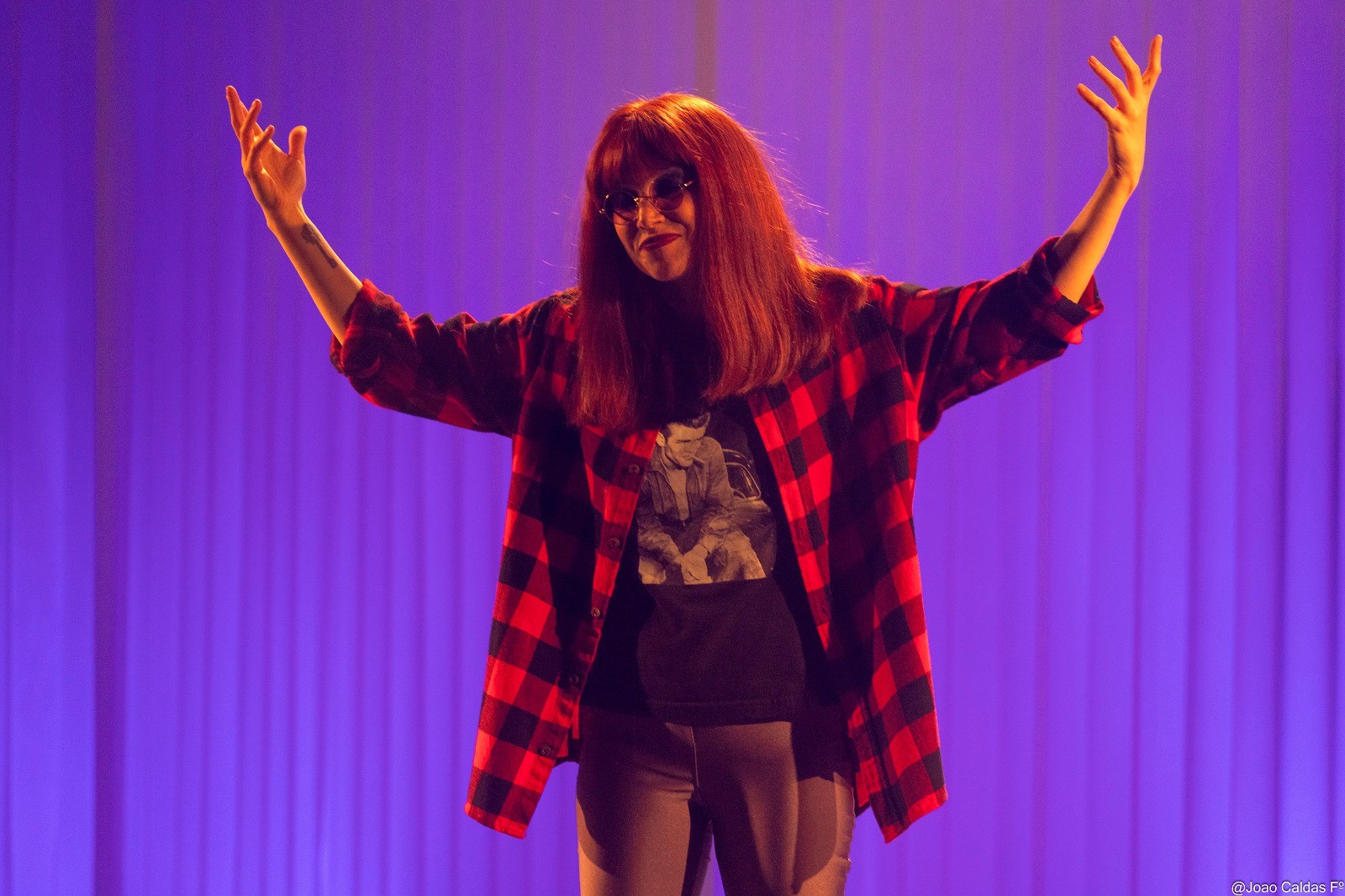 Mulher de cabelos ruivos e óculos escuros, vestindo camisa xadrez vermelha e preta sobre camiseta preta com estampa, braços erguidos com as mãos abertas, em um palco com iluminação roxa