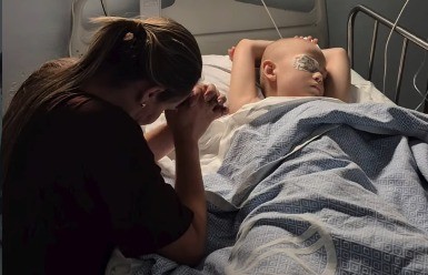 Mulher de cabelo preso, em perfil, com as mãos unidas em oração, ao lado de uma criança careca deitada em uma cama de hospital, com os olhos vendados e coberta por um lençol azul