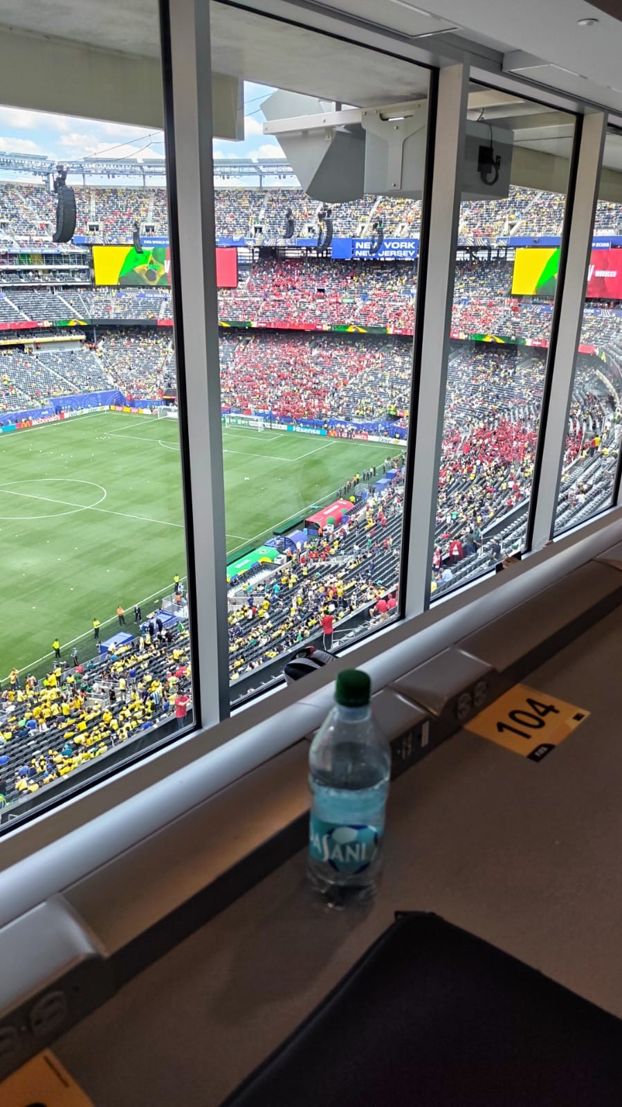 Vista de uma suíte em estádio lotado, com campo de futebol verde e torcedores nas arquibancadas. Uma garrafa de água e um número 104 em etiqueta amarela estão em uma bancada à frente da janela.