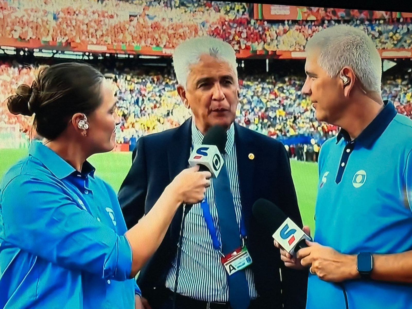 Três repórteres em campo, com um estádio lotado ao fundo. Uma mulher de camisa azul segura um microfone para um homem de terno e gravata, que fala. Outro homem de camisa polo azul, com um microfone na mão, observa a conversa