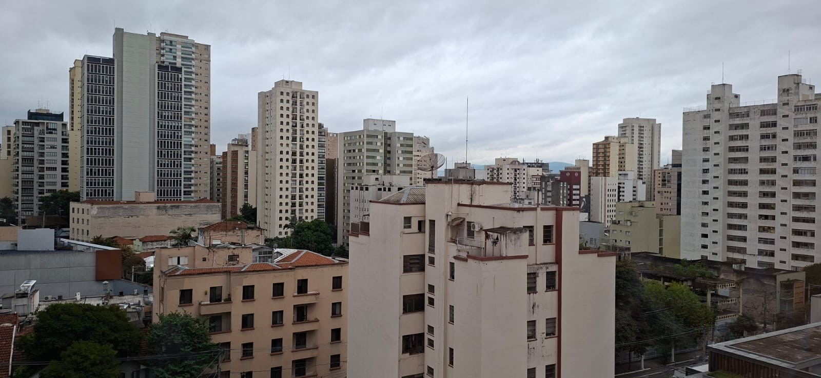 Vista aérea de uma cidade com muitos prédios residenciais e comerciais de diferentes alturas e cores claras, sob um céu nublado e cinzento. Algumas árvores verdes aparecem entre as construções, e antenas pontiagudas se destacam nos telhados. O cenário transmite uma atmosfera urbana e densa