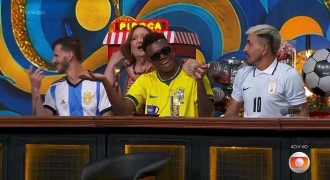 Quatro pessoas sentadas em um estúdio de TV. Um homem de camisa da Argentina sorri à esquerda. No centro, uma mulher de blusa colorida abraça um homem de óculos escuros e camisa do Brasil, que sorri. À direita, um homem de camisa do Uruguai com o número 10. Ao fundo, um cenário azul e amarelo com um letreiro Pipoca e uma bola de futebol