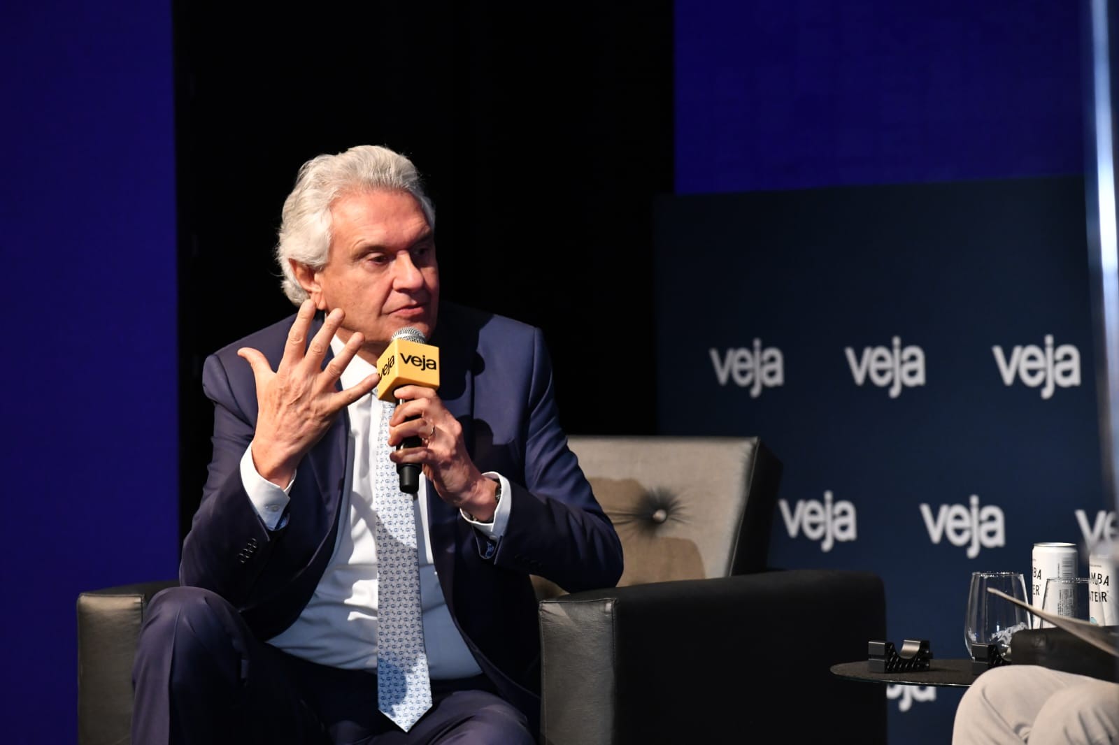 Homem de cabelos grisalhos, terno azul-marinho e gravata clara, sentado, gesticula com a mão esquerda enquanto segura um microfone amarelo com o logo Veja. Ao fundo, uma parede azul-escura com o logo Veja repetido em branco