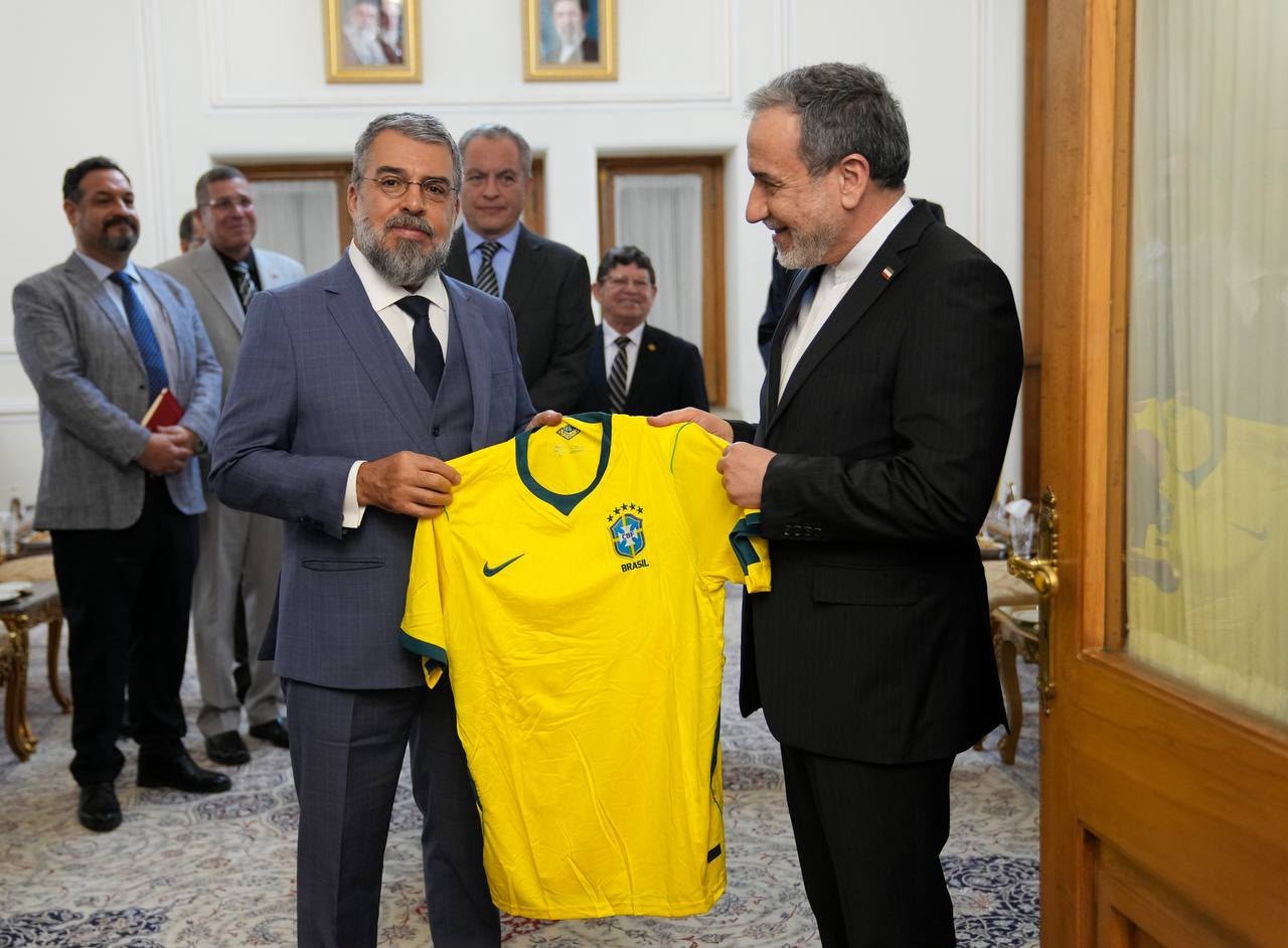 Dois homens de terno seguram uma camisa amarela da seleção brasileira de futebol, com o escudo da CBF. Outros homens de terno observam ao fundo, em um ambiente interno com quadros na parede