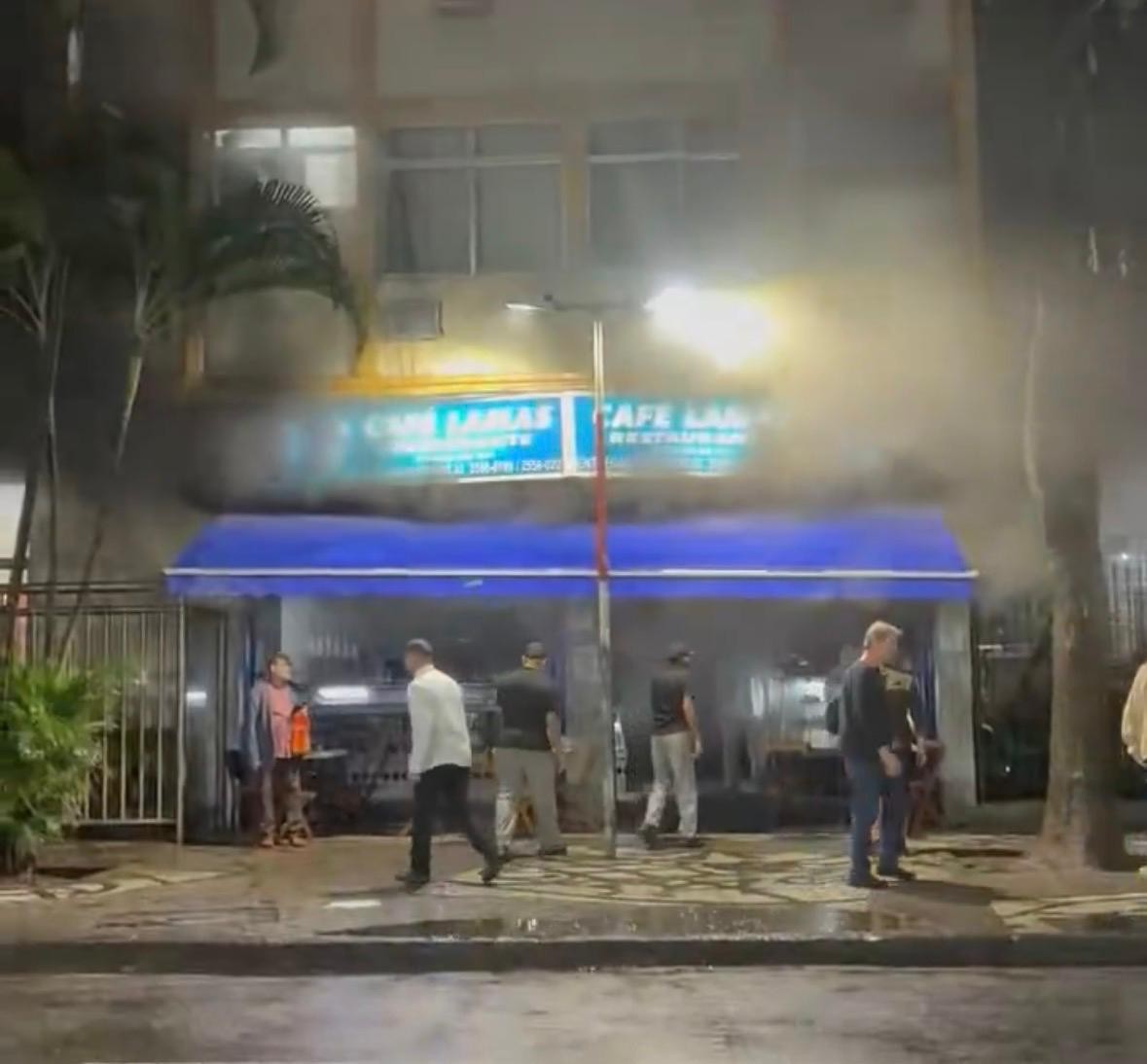 Fumaça densa saindo de um estabelecimento com toldo azul e letreiros luminosos Cafe Lamas, enquanto pessoas observam na calçada molhada à noite