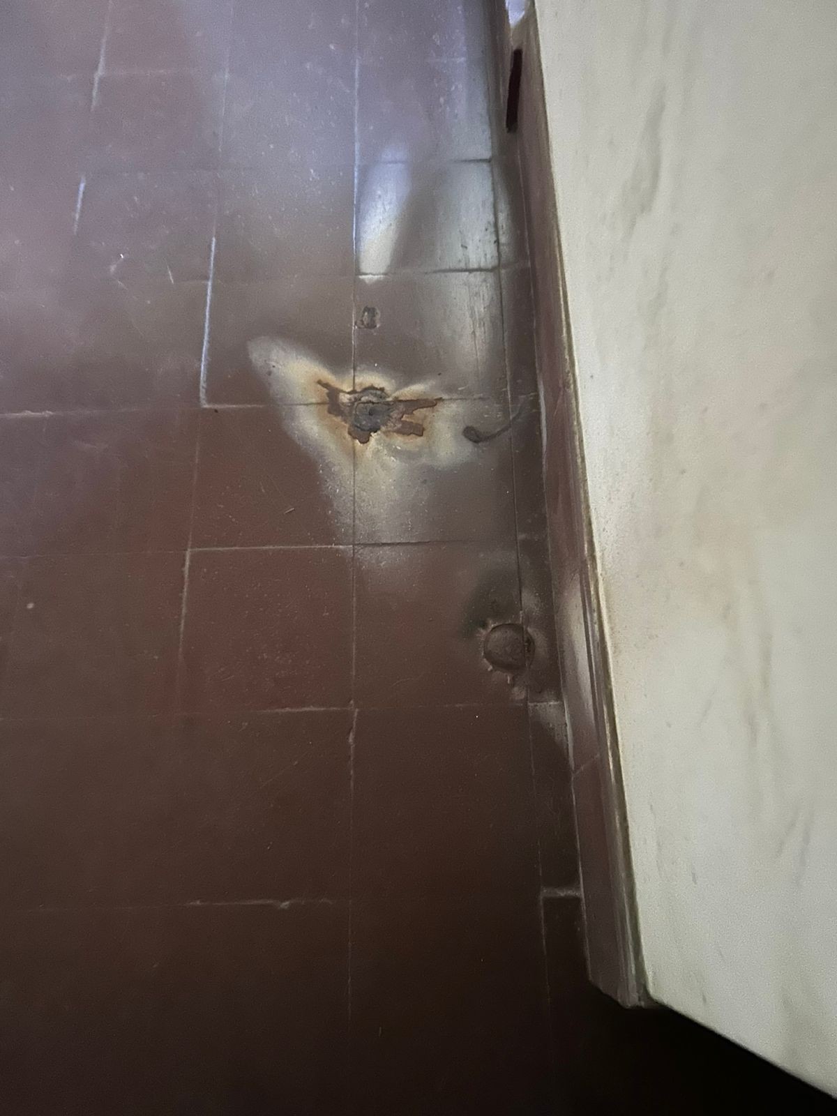 Piso de cerâmica marrom escuro com um buraco no centro, revelando terra e sujeira, e manchas brancas ao redor, indicando umidade ou vazamento