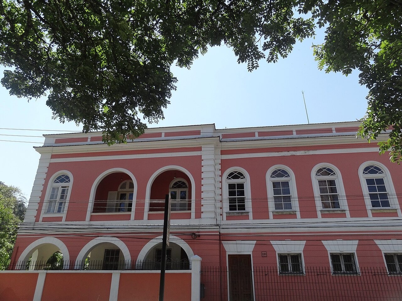 Fachada de um edifício histórico rosa com detalhes brancos, janelas arqueadas no andar superior e arcos no térreo, sob um céu azul claro com folhagem verde de árvores no topo