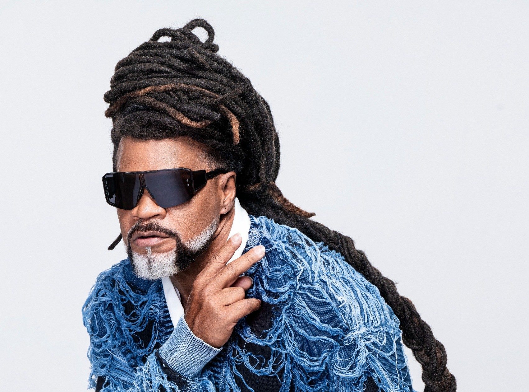Homem negro com dreadlocks longos, barba e bigode grisalhos, óculos escuros retangulares, vestindo um casaco azul desfiado e calça jeans, posa com a mão direita no queixo, em fundo branco