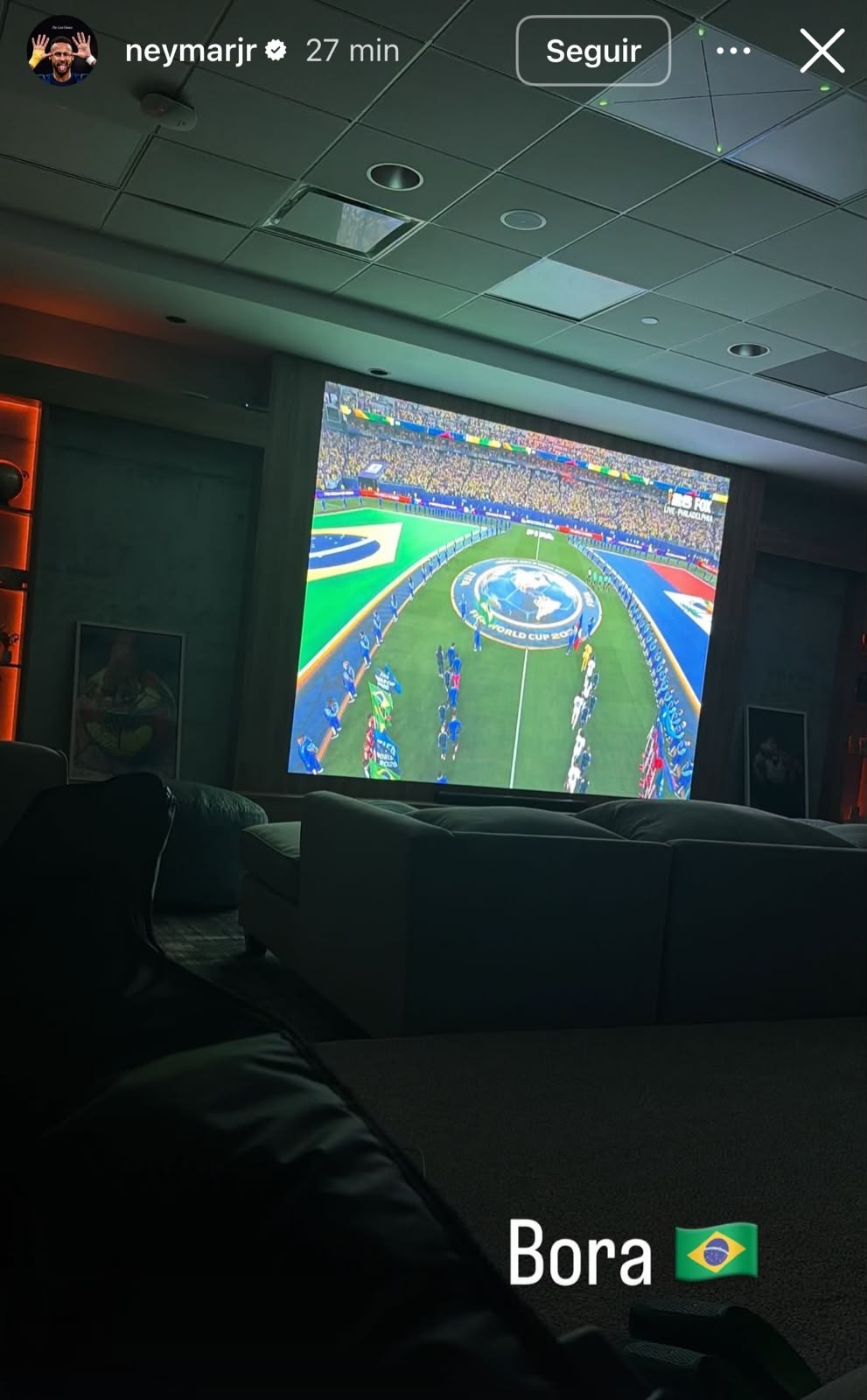 Pessoa assistindo a um jogo de futebol em uma tela grande de projeção, com o campo verde e a bandeira do Brasil visíveis, em uma sala escura com sofás confortáveis e iluminação ambiente
