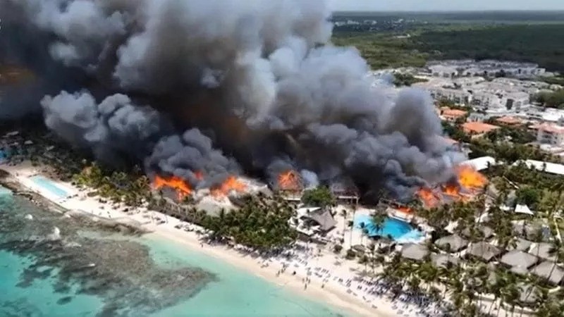 Vista aérea de um resort de praia com vários edifícios em chamas, liberando densa fumaça escura que se eleva ao céu. Pessoas aglomeradas na areia clara da praia, próxima ao mar azul-turquesa. Há piscinas azuis e coqueiros espalhados pelo local, com outras construções ao fundo. O céu está claro acima da fumaça