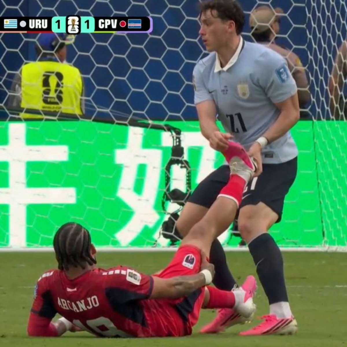 Dois jogadores de futebol em campo. Um, de camisa azul clara e shorts escuros, está em pé, segurando a perna do outro, que veste camisa vermelha com ARCANJO nas costas e está caído no gramado. O placar eletrônico acima mostra URU 1 1 CPV.