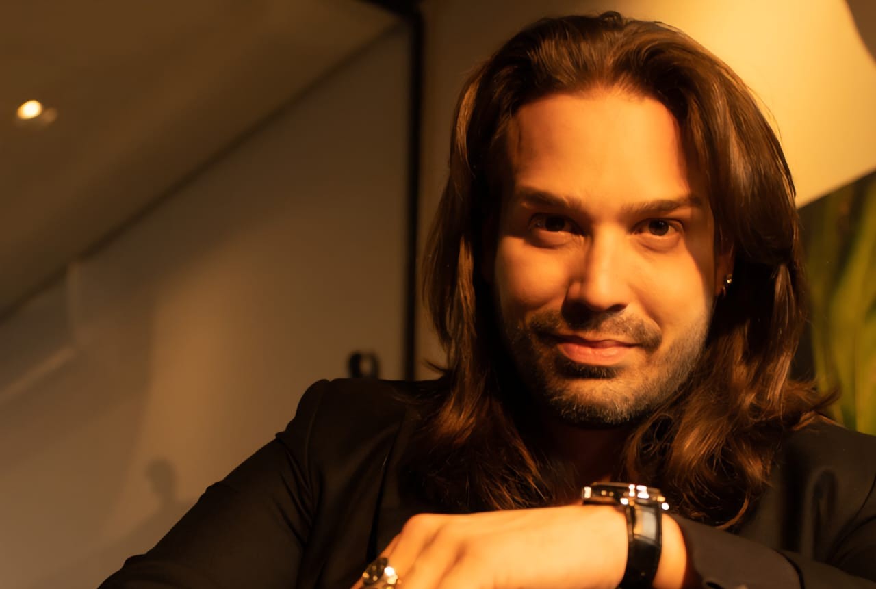 Homem de pele clara, cabelos castanhos longos e barba rala, sorri levemente para a câmera. Ele veste um blazer preto e usa um relógio de pulso escuro e anéis na mão direita, que está apoiada. A iluminação quente cria um ambiente aconchegante