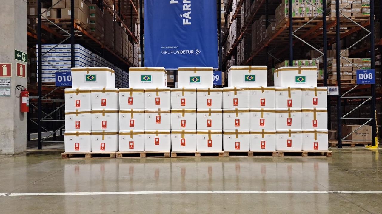 Caixas brancas empilhadas em paletes de madeira, com adesivos da bandeira do Brasil e selos vermelhos, em um armazém com prateleiras altas cheias de produtos e um banner azul ao fundo