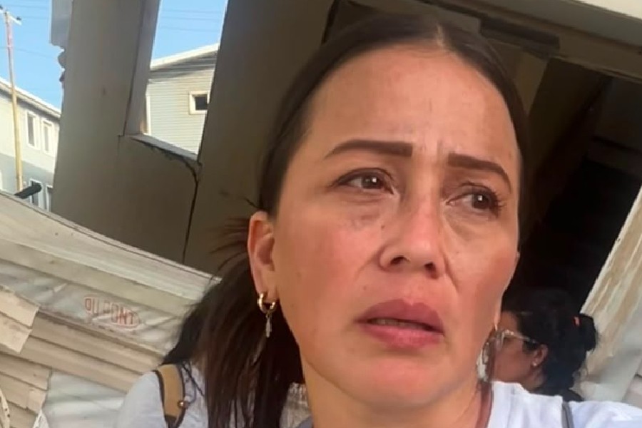 Mulher de pele morena clara, cabelo escuro preso, expressão de preocupação e olhos marejados, em primeiro plano. Ao fundo, estruturas de madeira e uma construção danificada, sugerindo cenário de destruição.