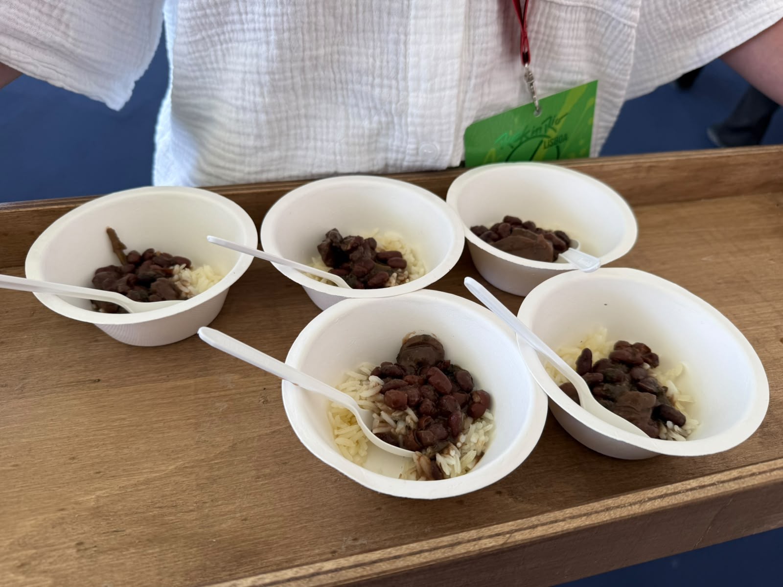 Cinco pequenas porções de arroz branco com feijão preto e colheres plásticas brancas, servidas em tigelas descartáveis brancas sobre uma bandeja de madeira. Uma pessoa com camisa branca e crachá verde segura a bandeja