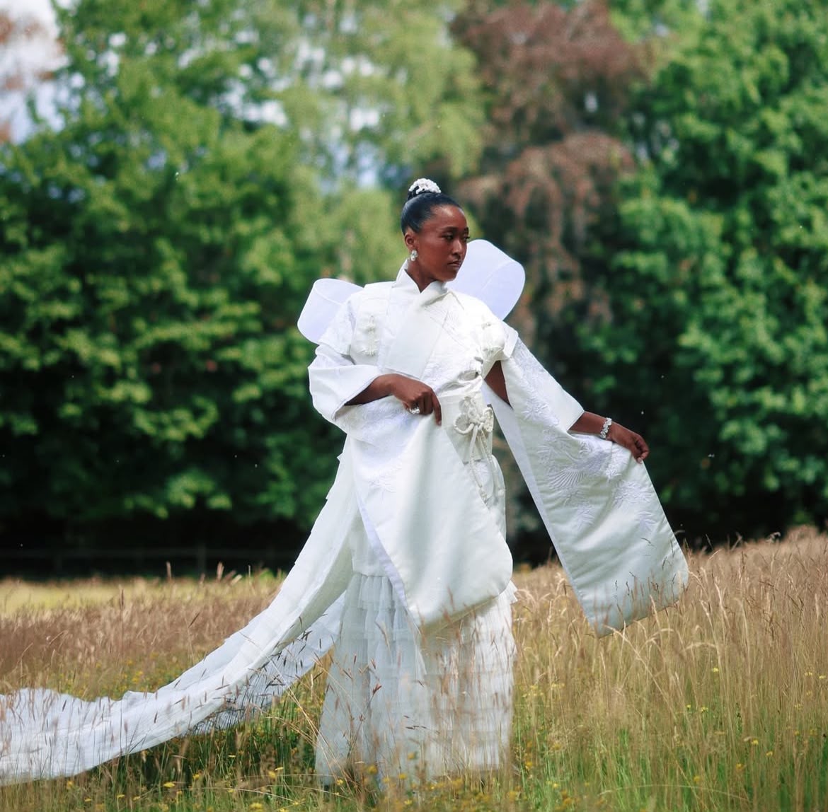 Mulher negra em vestido branco longo e esvoaçante, com asas brancas nas costas, em pé num campo de grama alta e seca, com árvores verdes ao fundo