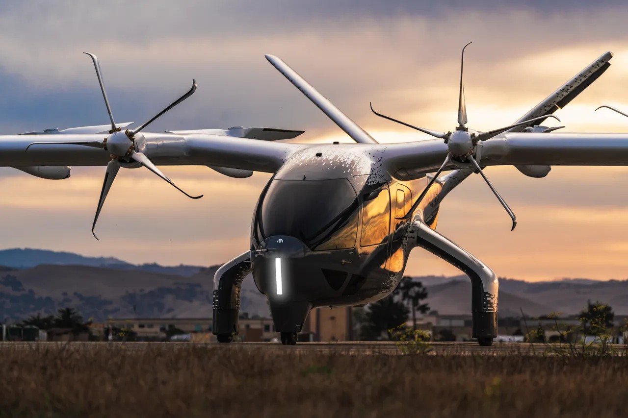 Aeronave preta e cinza com quatro hélices em cada asa, parada em uma pista de terra com montanhas ao fundo e céu alaranjado no pôr do sol