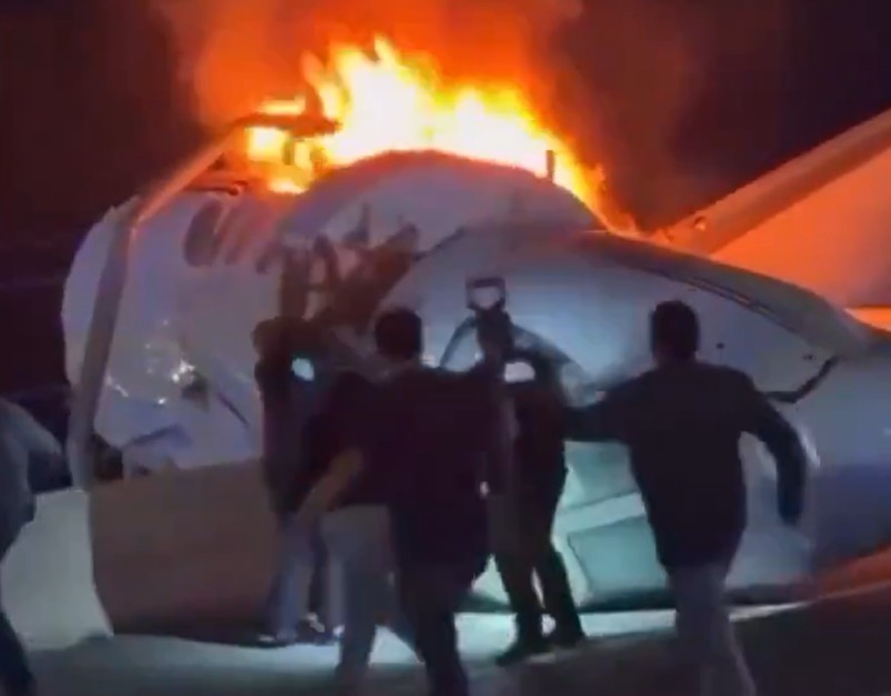 Pessoas correm perto de um avião branco em chamas, com grandes labaredas laranja e fumaça escura saindo da fuselagem, em um cenário noturno