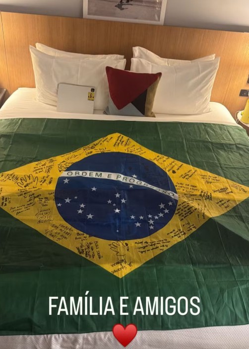 Cama de hotel com lençóis brancos, travesseiros e uma bandeira do Brasil estendida sobre o edredom. A bandeira tem a parte amarela coberta por diversas assinaturas e mensagens. Um tablet e uma almofada decorativa estão sobre os travesseiros. Na parte inferior da imagem, lê-se FAMÍLIA E AMIGOS com um coração vermelho abaixo