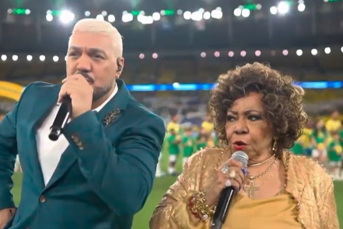 Cantores Belo, com cabelo platinado e terno verde-azulado, e Alcione, com cabelo cacheado e casaco dourado, cantam em um palco de estádio iluminado, segurando microfones