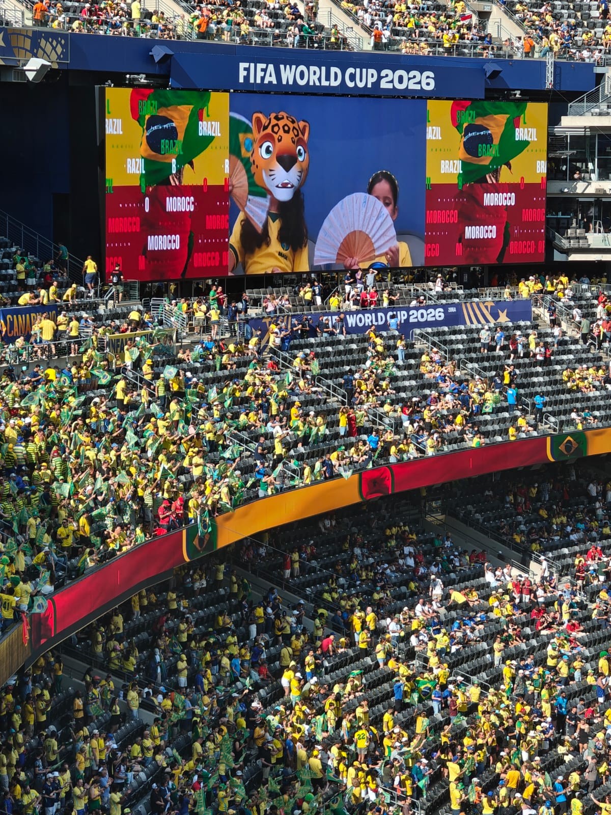 Torcida em estádio lotado, com pessoas vestindo amarelo e verde, e um telão exibindo FIFA WORLD CUP 2026 e mascotes com leques