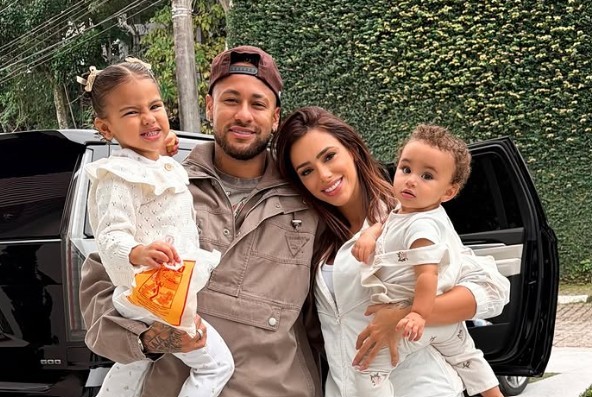 Neymar, Bruna Biancardi e suas duas filhas posam sorridentes em frente a um carro preto e um muro verde. Neymar usa boné e jaqueta marrom, segurando a filha mais velha, que veste um suéter branco e segura um pacote laranja. Bruna, de macacão branco, segura a filha mais nova, de macacão claro.