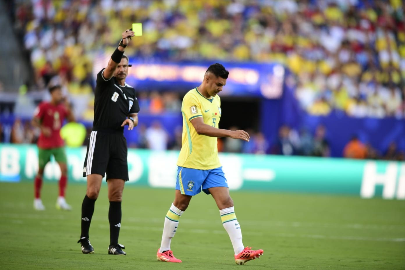 Um árbitro de futebol, vestindo uniforme preto, levanta um cartão amarelo para um jogador brasileiro, que usa camisa amarela e shorts azuis, caminhando com expressão séria em campo verde. Torcedores desfocados e coloridos preenchem as arquibancadas ao fundo