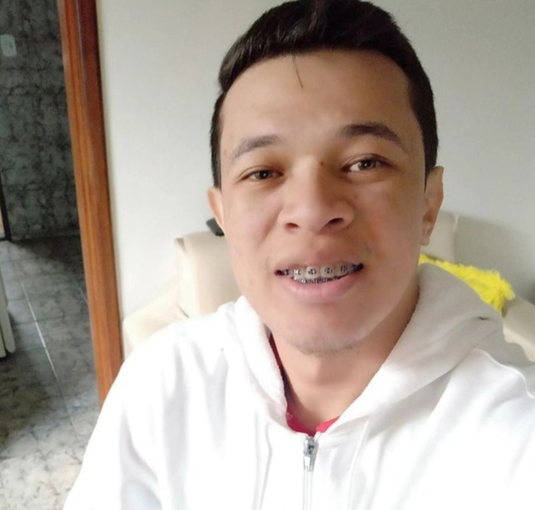 Homem jovem de pele clara, cabelo escuro penteado para cima, usando aparelho ortodôntico e um moletom branco com zíper, sorrindo levemente para a câmera em um ambiente interno com paredes claras e um sofá bege ao fundo